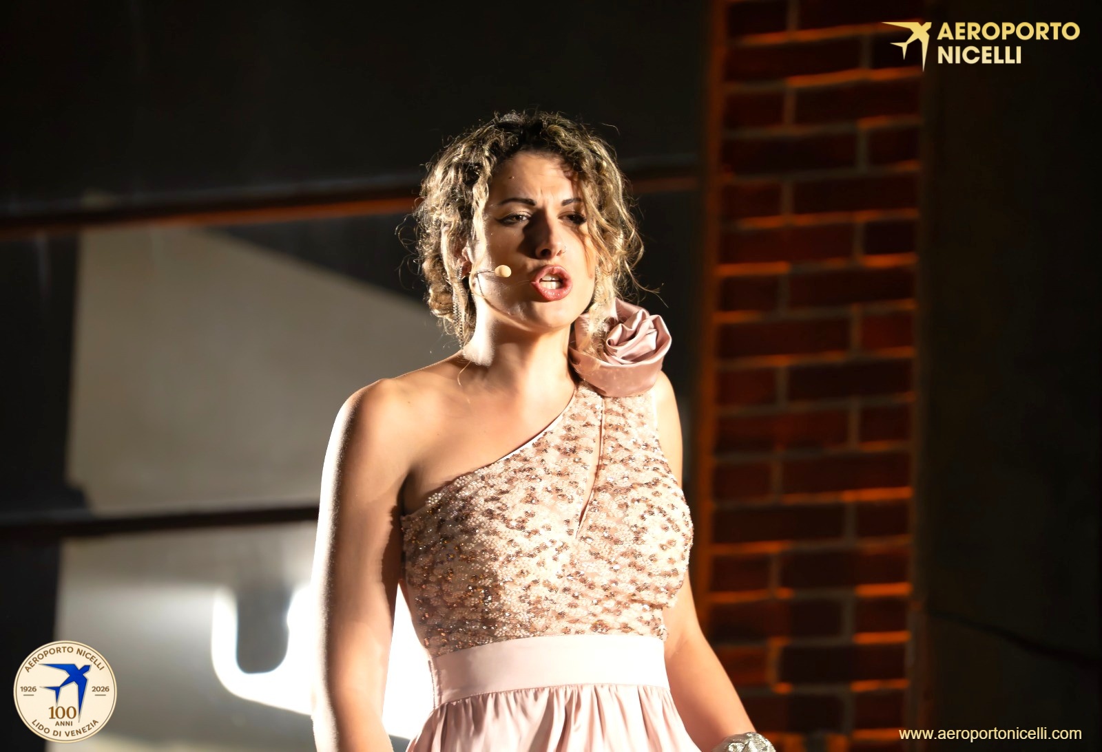 Una donna che canta sul palco, indossando un abito rosa con decorazioni scintillanti e un fiore sulla spalla.
