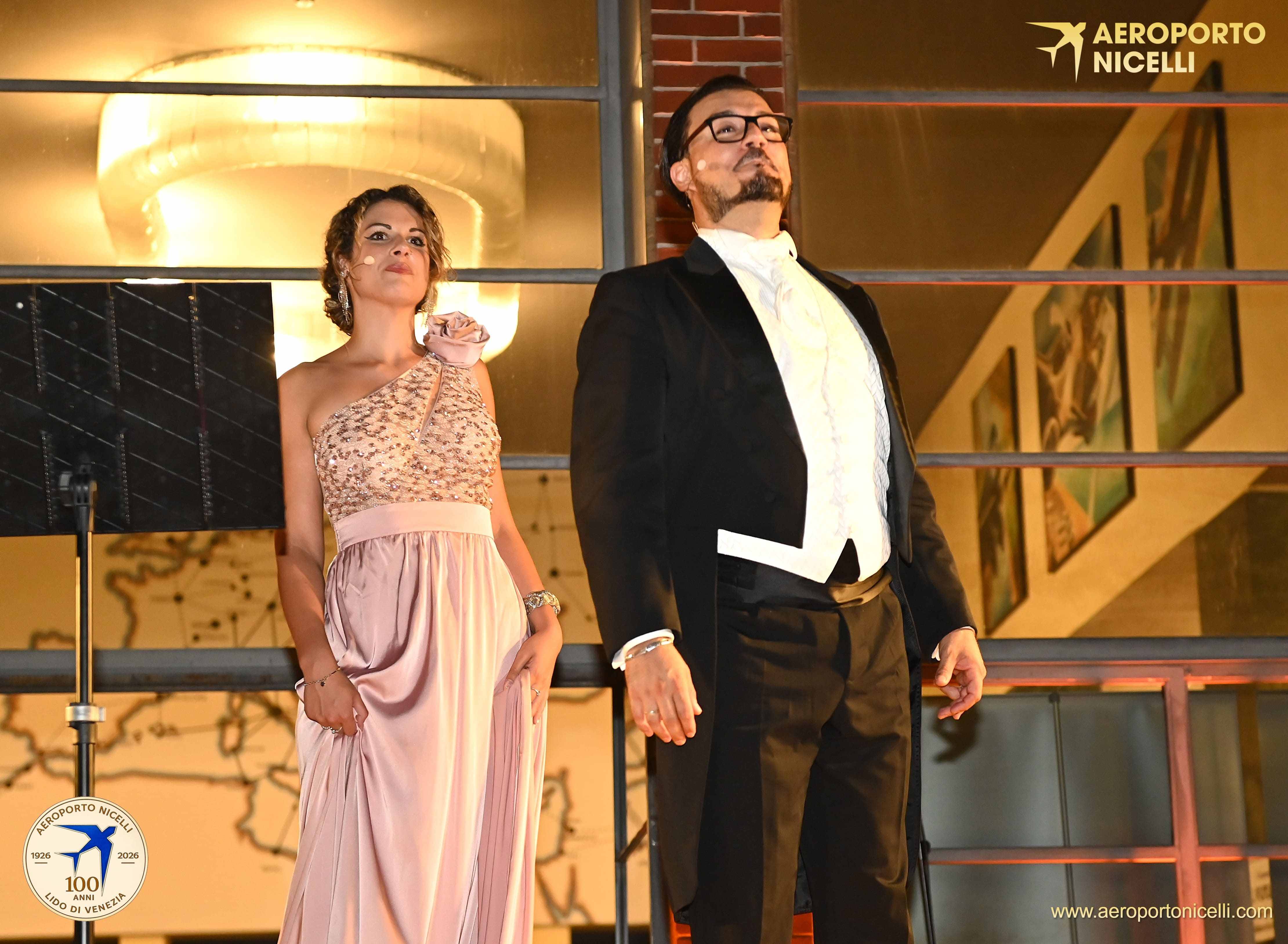 Due performer in abbigliamento elegante sul palco, con un lampadario sullo sfondo e opere d'arte sulle pareti.