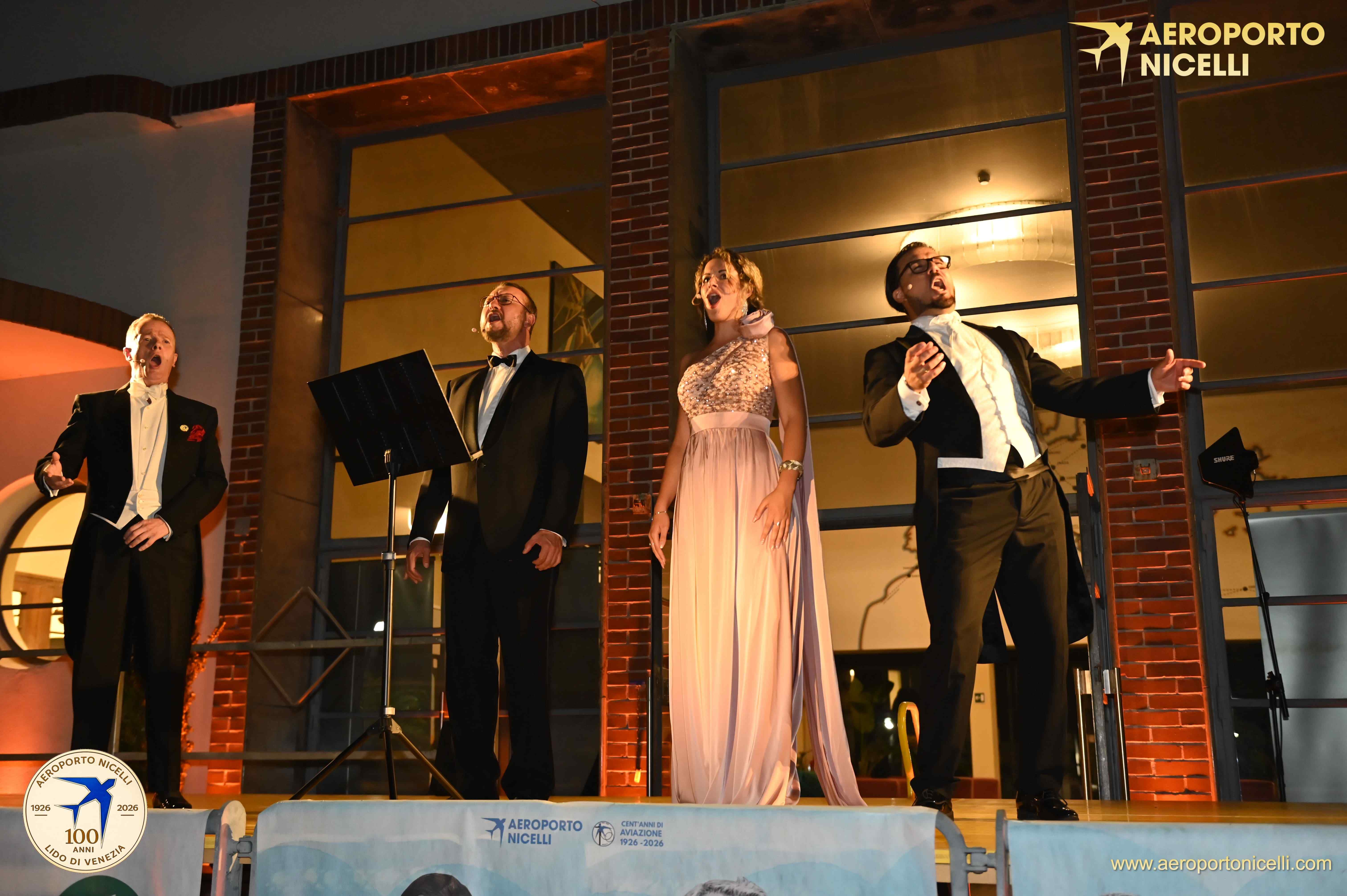 Un gruppo di quattro cantanti si esibisce su un palco all'aperto, indossando abiti eleganti. Due uomini e una donna sono in primo piano mentre cantano con passione, illuminati da luci soffuse.