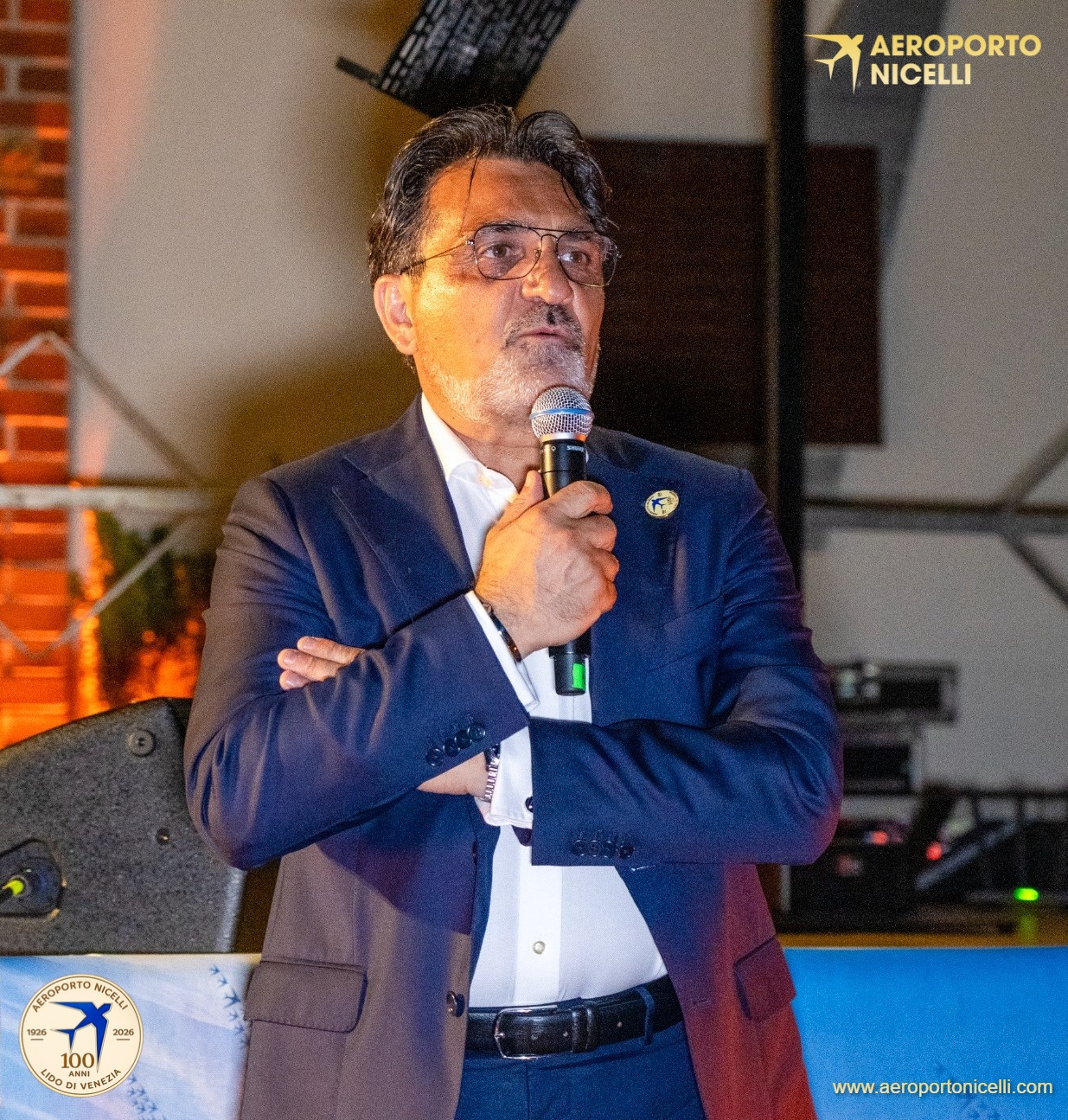 Uomo in abito scuro parla al microfono durante un evento all'aperto, con sfondo di luci e attrezzature audio.