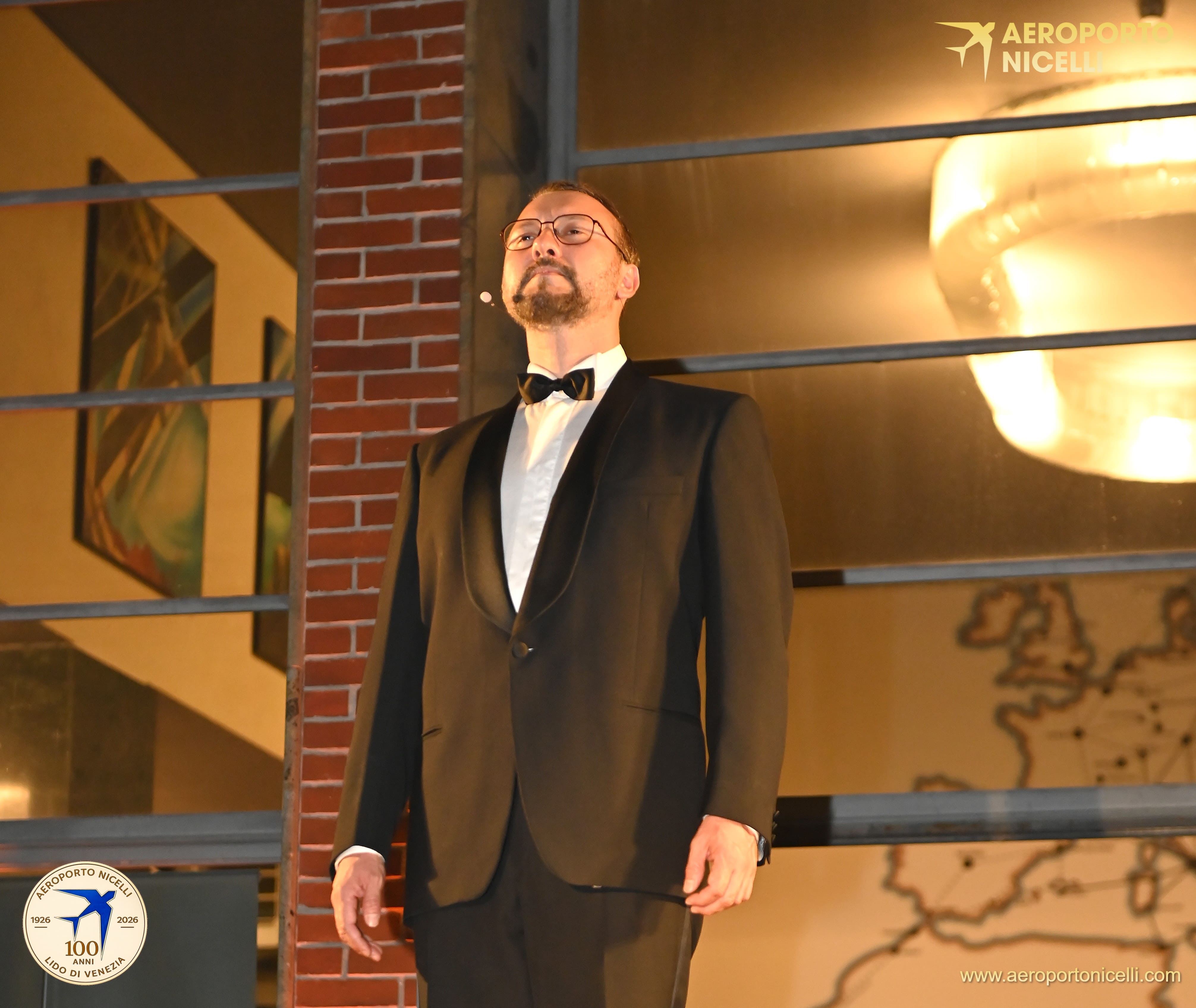 Un uomo in smoking con occhiali, in piedi su un palco, che sembra pronto a esibirsi, con un'illuminazione calda sullo sfondo.