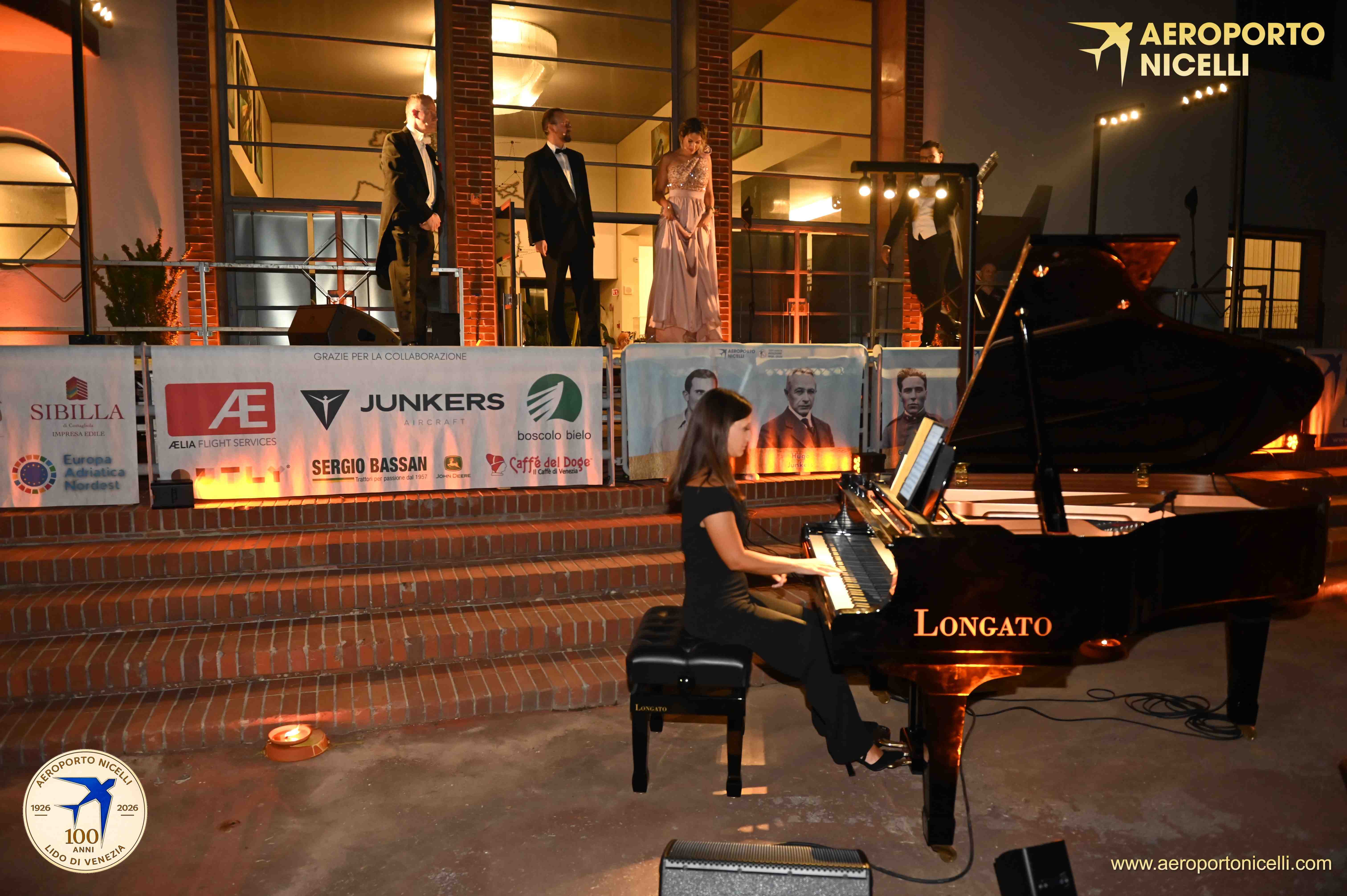 Una pianista suona un pianoforte a coda durante un evento, mentre sul palco si esibiscono cantanti. Sullo sfondo, vari sponsor sono esposti su un pannello.