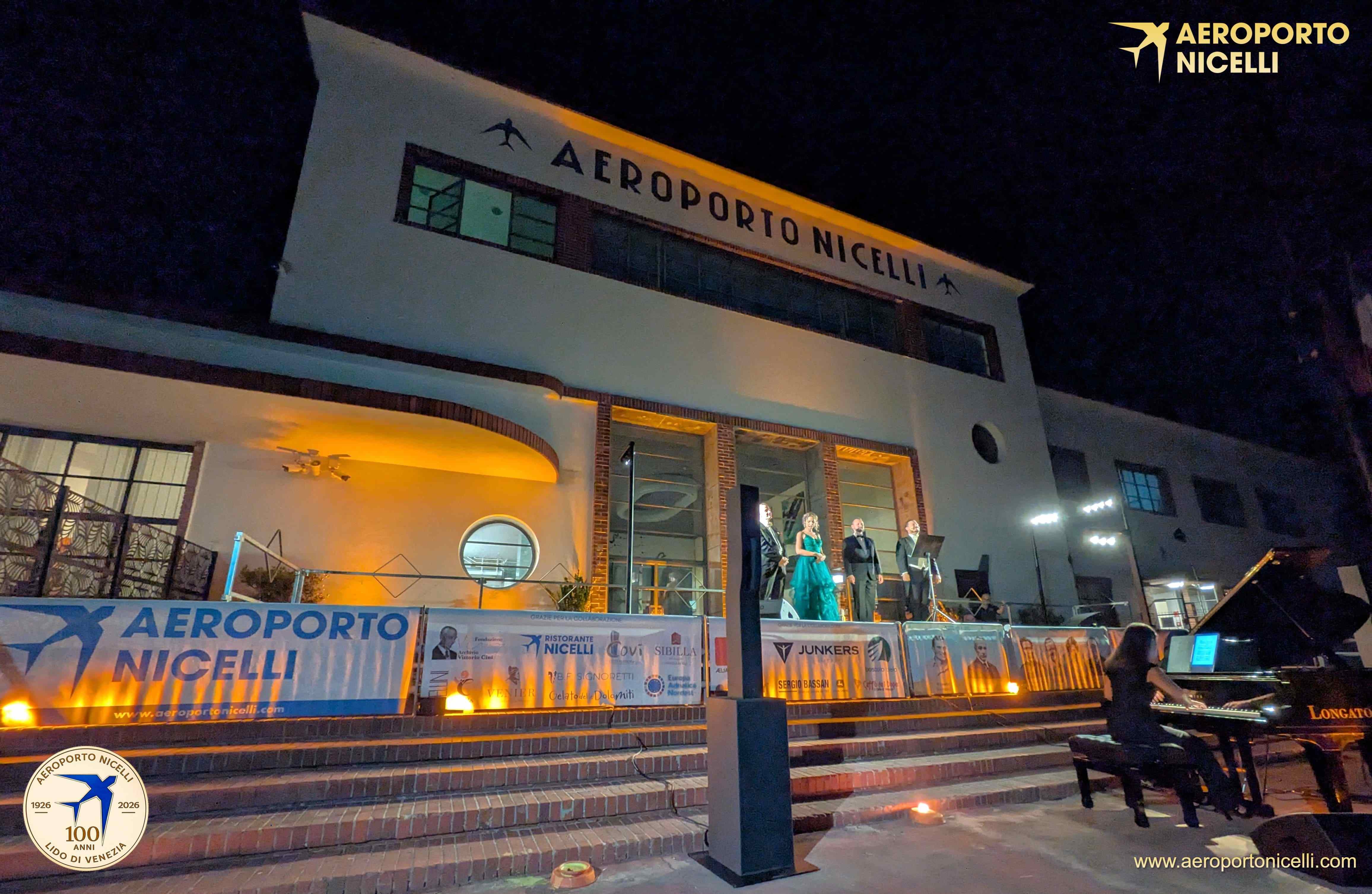 Un evento musicale notturno presso l'Aeroporto Nicelli, con artisti che si esibiscono sul palco e un pianista che suona.